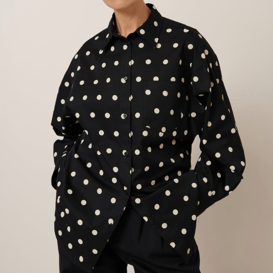 A woman modeling a long-sleeved shirt with a large-scale polka dot pattern in black and forest green colors, with a collar and button cuffs.