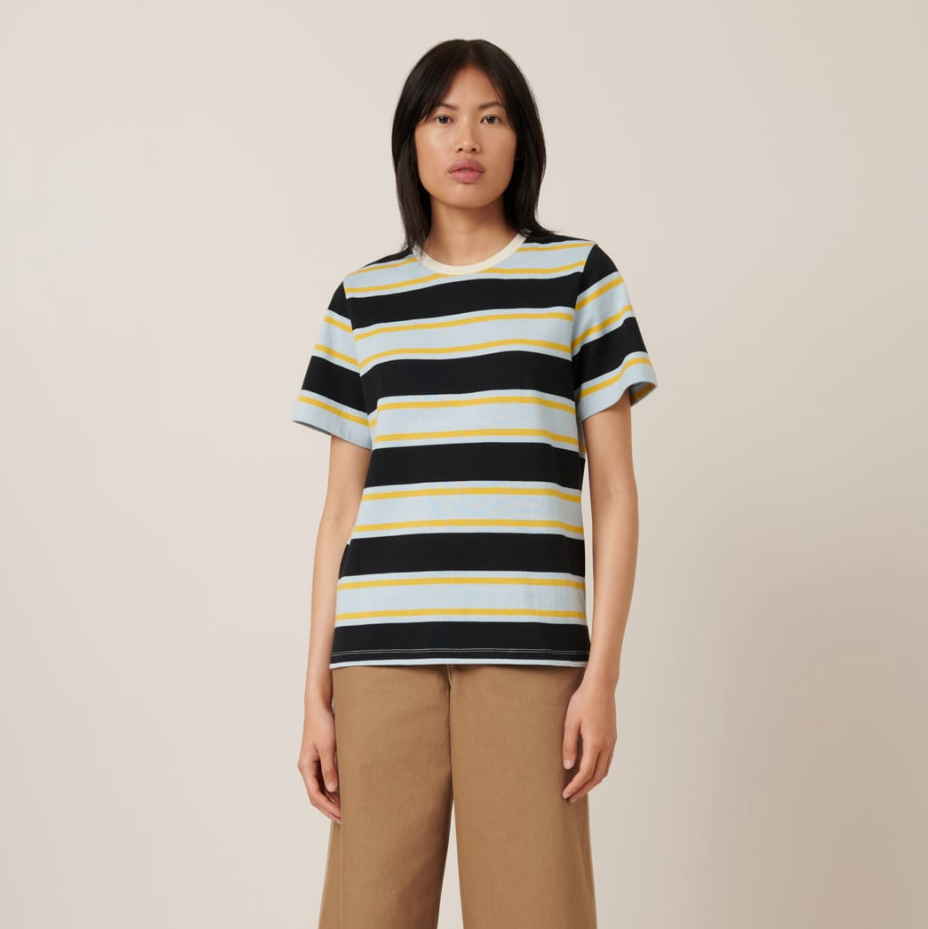 A person standing against a neutral background, wearing a striped short sleeve shirt with a ribbed crew neck and long pants.