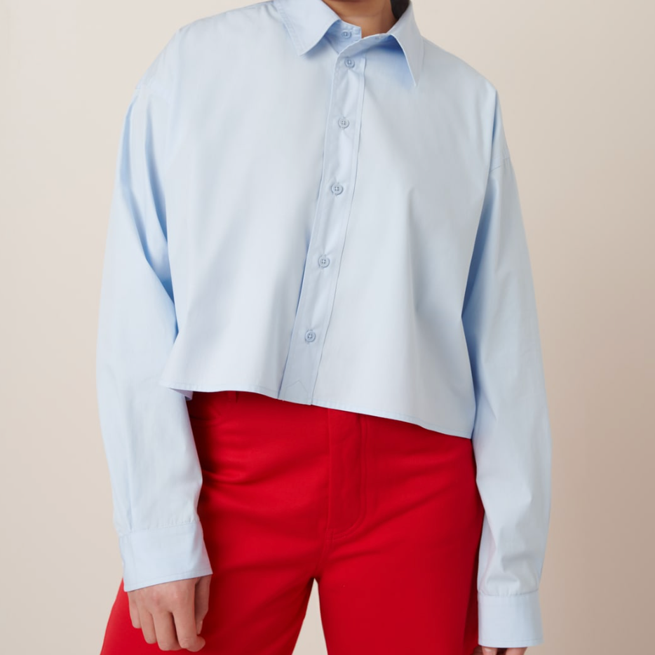 A woman wearing a sky blue boxy, cropped Lauren Shirt with a classic shirt collar and pleated cuffs, paired with red trousers.