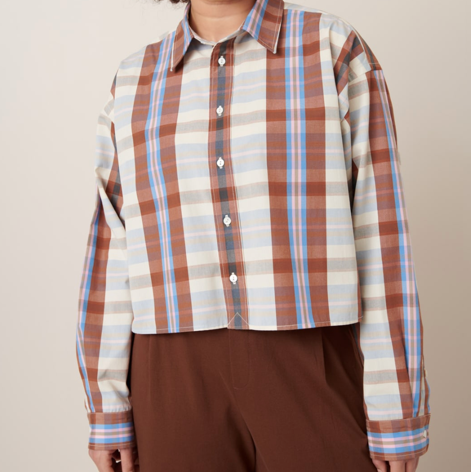 A woman is wearing a boxy, cropped style shirt with a classic collar and pleated cuffs, buttoned at the top with a pattern of sky blue and brown stripes.