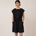 A model wearing a black knee-length dress with oversized front pockets, short sleeves, and large buttons. The dress has a drawcord at the waist and appears to be made of cotton.
