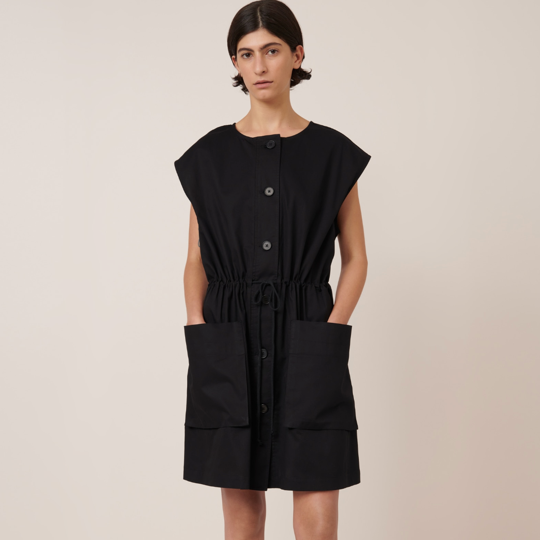 A model wearing a black knee-length dress with oversized front pockets, short sleeves, and large buttons. The dress has a drawcord at the waist and appears to be made of cotton.