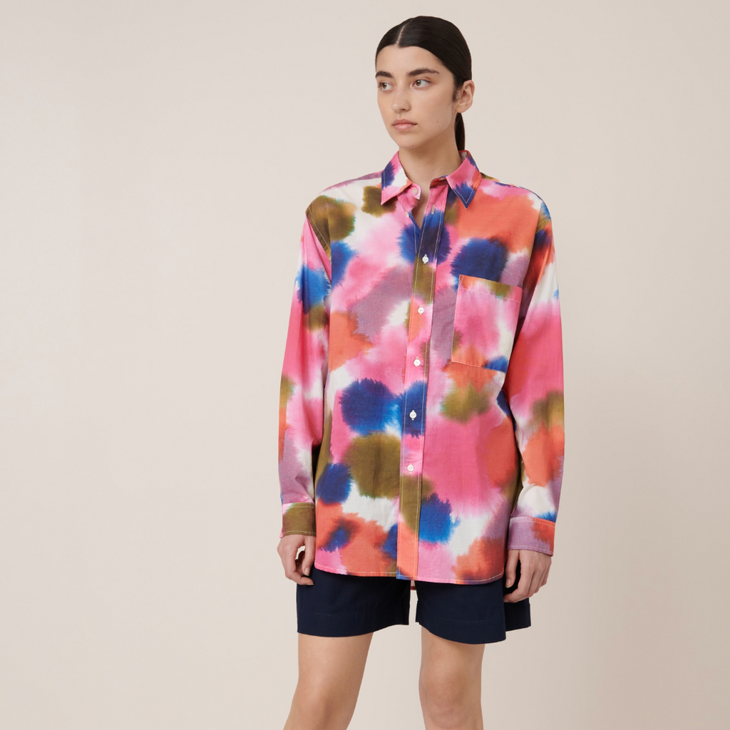 A model wearing an oversized multicolored shirt with a classic collar and long sleeves, paired with dark shorts and black shoes.
