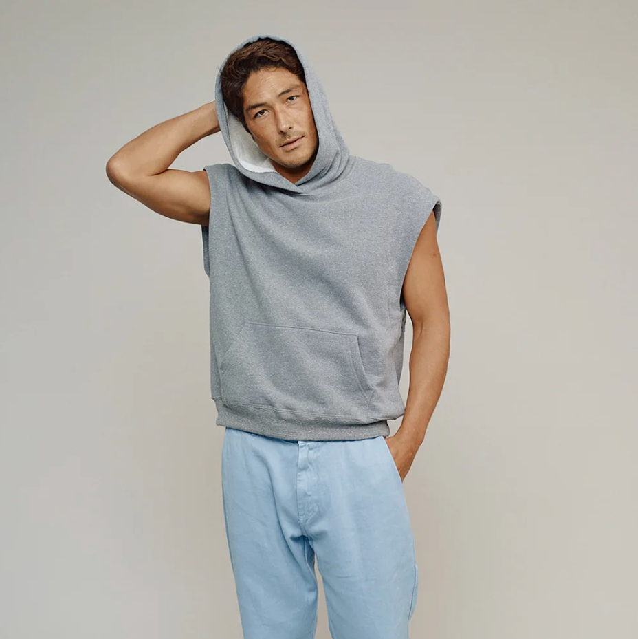 Man wearing a sleeveless gray hoodie and light blue jeans on a plain background