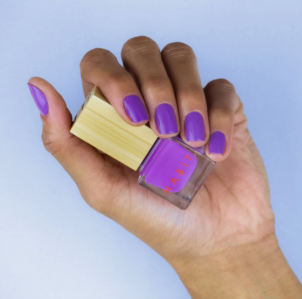 Hand holding a bottle of purple nail polish with a matching color swatch on a light blue background
