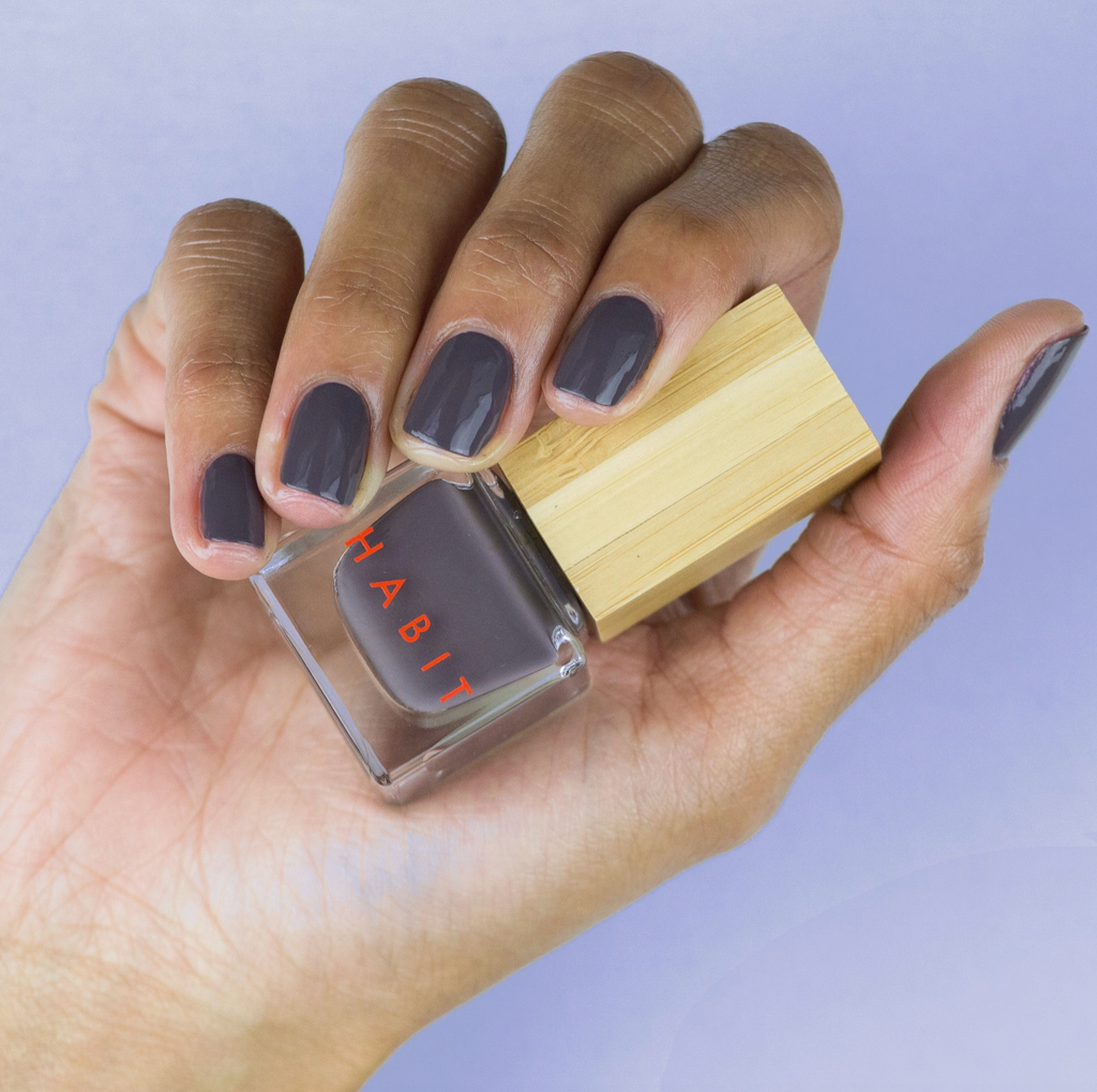Hand holding a bottle of nail polish with a wooden cap against a light purple background