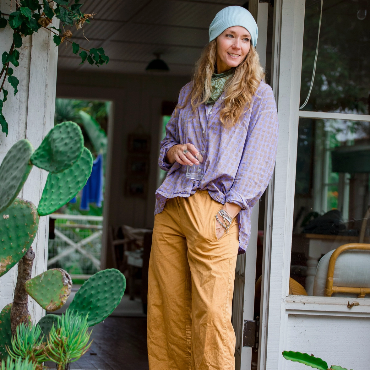 Woman standing on a wooden porch with plants and a white building in the background
