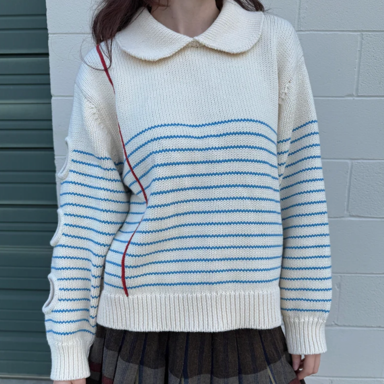 Person wearing a white sweater with blue and red stripes against a gray wall.