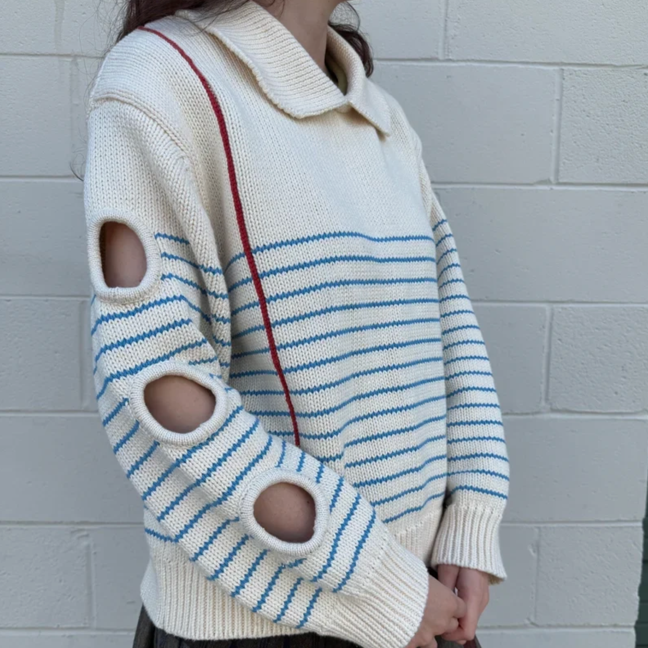 Person wearing a white sweater with blue stripes and cut-out sleeves against a gray brick wall.